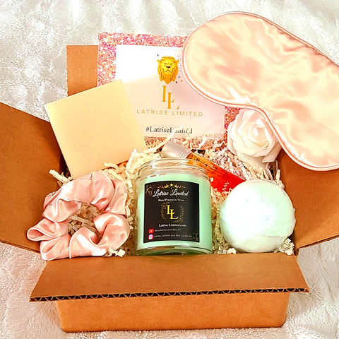 This image contains a Sage and Vanilla 6oz candle, an unscented soap, an eucalyptus bath bomb, a pain essential oil blend, a pink silk eye mask, and a pink silk scrunchy. The items are all inside of a box and the background is white. 