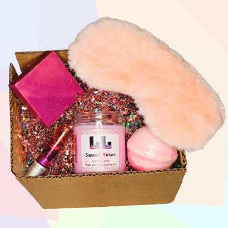 This image contains a Sage and Vanilla 6oz candle, an unscented soap, an eucalyptus bath bomb, a pain essential oil blend, a pink silk eye mask, and a pink silk scrunchy. The items are all inside of a box and the background is white. 